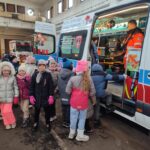 Grupa dzieci w zimowych ubraniach stoi przy karetce pogotowia, do której wchodzi sanitariusz.