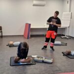 Dziewczyna ćwiczy resuscytację na manekinie treningowym, podczas gdy instruktor obserwuje ją w klasie.