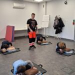 Mężczyzna instruuje dzieci ćwiczące resuscytację krążeniowo-oddechową na manekinach w klasie.