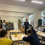 Nauczyciel stoi i przemawia do uczniów siedzących przy stolikach w klasie z książkami i roślinami.