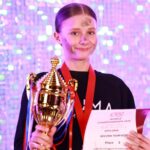 Uśmiechnięta dziewczyna trzymająca trofeum i certyfikat, nosząca medal, stojąca przed błyszczącym tłem.