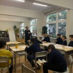 Nauczyciel przemawia do uczniów siedzących przy biurkach w jasnej klasie z dużymi oknami i roślinami.