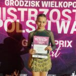 Dziewczyna w błyszczącym kostiumie tanecznym trzyma certyfikat, stojąc przed tłem zawodów.