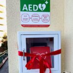 Defibrylator w obudowie naściennej z czerwoną wstążką i napisem instruktażowym powyżej.