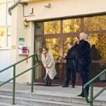 Trzy kobiety stoją przed wejściem do budynku w pobliżu schodów i poręczy, odbijając się w szklanych drzwiach.