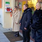 Trzy osoby stoją przy wejściu do budynku; jedna kobieta mówi do mikrofonu w pobliżu zamontowanego na ścianie defibrylatora.