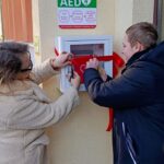 Dwie osoby przecinają czerwoną wstęgę na zewnętrznej skrzynce AED (defibrylatora) zamontowanej na ścianie.