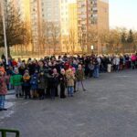 Duża grupa ludzi, w tym dzieci, gromadzi się na zewnątrz w pobliżu budynku w zimny dzień.