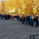 Duża grupa ludzi stoi w kolejce na zewnątrz na utwardzonym terenie, z drzewami i budynkami w tle.