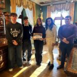 Pięć osób stoi w oświetlonym słońcem, ozdobnym pokoju; jedna osoba w środku trzyma oprawione zdjęcie.