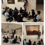 Dzieci siedzą na podłodze muzeum, słuchając przewodnika omawiającego obrazy i wystawy sztuki na ścianach.