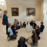 Grupa dzieci siedzi wokół marmurowej rzeźby w muzeum sztuki i słucha przemawiającej kobiety.