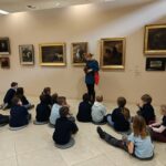 Grupa dzieci siedzi na podłodze i słucha kobiety w galerii sztuki. Obrazy wiszą z tyłu.