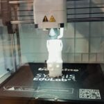 Drukarka 3D w akcji, drukująca biały obiekt na swojej platformie wewnątrz przezroczystej obudowy.