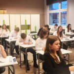 Uczniowie w klasie siedzą przy biurkach, skupieni na pisaniu lub czytaniu prac podczas testu.