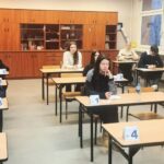 Uczniowie siedzą przy osobnych biurkach w klasie, każdy z ponumerowaną kartą, zdając test.