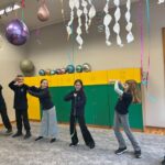 Pięcioro dzieci tańczy w kolorowej klasie udekorowanej balonami, serpentynami i wiszącymi wstążkami.