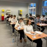 Uczniowie siedzą przy osobnych biurkach w klasie, zdając egzamin z papierami i butelkami wody przed sobą.