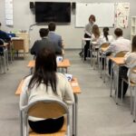 Uczniowie siedzą przy pojedynczych biurkach w klasie i zdają egzamin pod nadzorem nauczyciela.