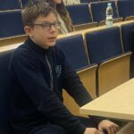 Chłopiec w okularach siedzi w sali wykładowej z innymi studentami w tle.