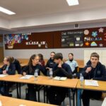 Uczniowie w mundurkach siedzą przy biurkach w klasie, rozmawiając i przygotowując się do dyskusji.