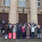 Grupa dzieci w zimowych ubraniach stoi na kamiennych schodach, unosząc ręce przed dużym budynkiem.