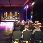 Dzieci siedzą w rzędach krzeseł naprzeciwko sceny ze statywami muzycznymi i różowymi balonami na podłodze.