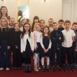 Grupa dzieci i kobieta stoją razem w pomieszczeniu na czerwonym dywanie, twarzą do kamery.