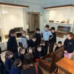 Grupa dzieci gromadzi się wokół ekspozycji muzealnej, słuchając wyjaśnień kobiety.