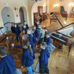 Grupa dzieci stoi w sali muzealnej wypełnionej różnymi zabytkowymi fortepianami i drewnianymi podłogami.