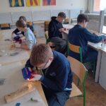 Czterech uczniów w klasie używa narzędzi do pracy nad projektami z drewna; znak ART na ścianie.