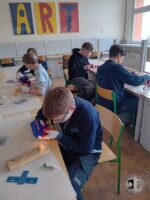 Czterech uczniów w klasie używa narzędzi do pracy nad projektami z drewna; znak ART na ścianie.