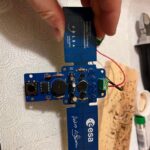 Ręka trzyma płytkę drukowaną ESA w kształcie krzyża na ręczniku papierowym z narzędziami w pobliżu.