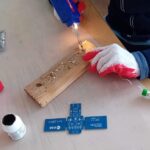 Osoba wierci otwory w drewnianej desce w pobliżu niebieskiej płytki drukowanej na jasnym stole.