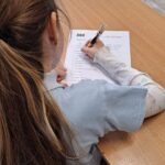 Dziewczyna z długimi włosami pisze odpowiedzi na arkuszu przy drewnianym biurku w klasie.