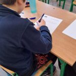 Uczeń siedzi przy biurku, pisząc na kartce, z długopisami i butelką wody w pobliżu.