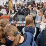 Duża grupa ludzi zebrała się w środku wokół stołów z kolorowymi przedmiotami na wystawie.