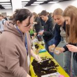 Uczniowie gromadzą się wokół stołu, sadząc nasiona w glebie na zatłoczonym wydarzeniu lub targach.