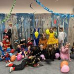 Dzieci w kolorowych kostiumach pozują z balonami i serpentynami w udekorowanej klasie na przyjęcie.