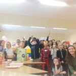 Grupa dzieci i dorosłych w kostiumach pozuje i uśmiecha się w udekorowanej sali lekcyjnej.