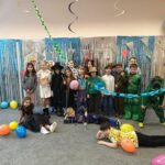 Grupa dzieci w kostiumach pozuje razem na kolorowej, świątecznej imprezie z balonami i serpentynami.