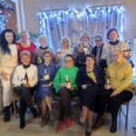 Dwanaście kobiet pozuje w pomieszczeniach, trzymając udekorowane kubki, a za nimi znajdują się świąteczne lampki i zimowe dekoracje.