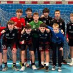 Jedenastu chłopców w strojach sportowych pozuje przed bramką do piłki ręcznej na krytym boisku.