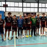 Drużyna piłki ręcznej chłopców w strojach pozująca z medalami na sali gimnastycznej, za nimi dwa niebieskie banery.