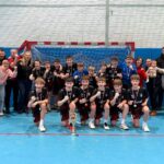 Młodzieżowa drużyna piłkarska pozuje z trofeum przed bramką, otoczona dopingującymi kibicami.