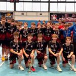 Chłopięca drużyna sportowa pozuje w pomieszczeniu z medalami i trofeum, otoczona przez dwóch dorosłych w czarnych garniturach.