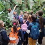 Grupa dzieci z plecakami słucha przewodnika w bujnym, krytym ogrodzie botanicznym.