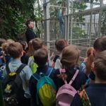 Dzieci z plecakami obserwują opiekuna zoo i dużego ptaka wewnątrz woliery lub wybiegu zoo.