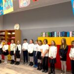 Dzieci stoją w rzędzie w klasie, ubrane w białe bluzki, a nad nimi wiszą kolorowe dzieła sztuki.