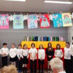 Grupa dzieci i dorosły stoją w klasie pod kolorowymi rysunkami zimowych płaszczy.